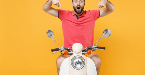 Jongen met baard zit op scooter en maakt een gebaar met beide wijsvingers naar beneden naar de scooter