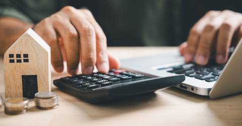 Een man is aan het rekenen op een rekenmachine met op de voorgrond een huisje en munten